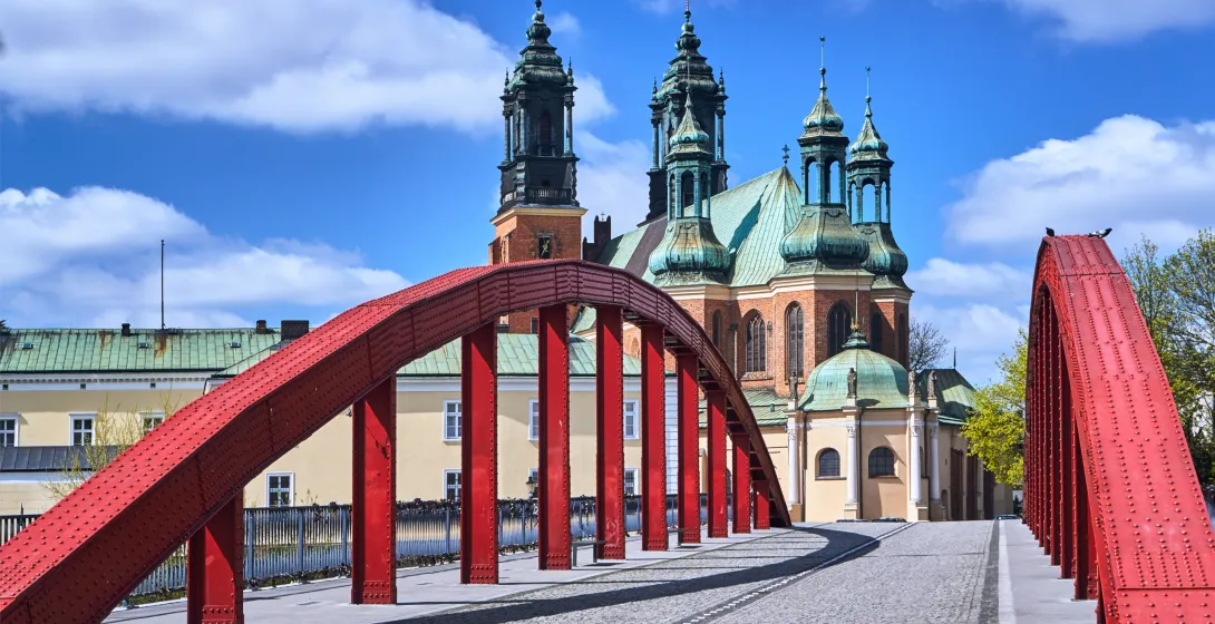 Red steel arch bridge leading to Poznań Cathedral with green copper domes under a blue sky with white clouds.
