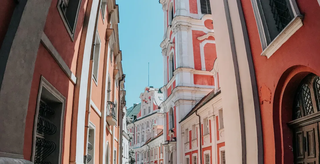 Cobblestone street between historic buildings with red and white facades, leading to a baroque church tower with a green dome