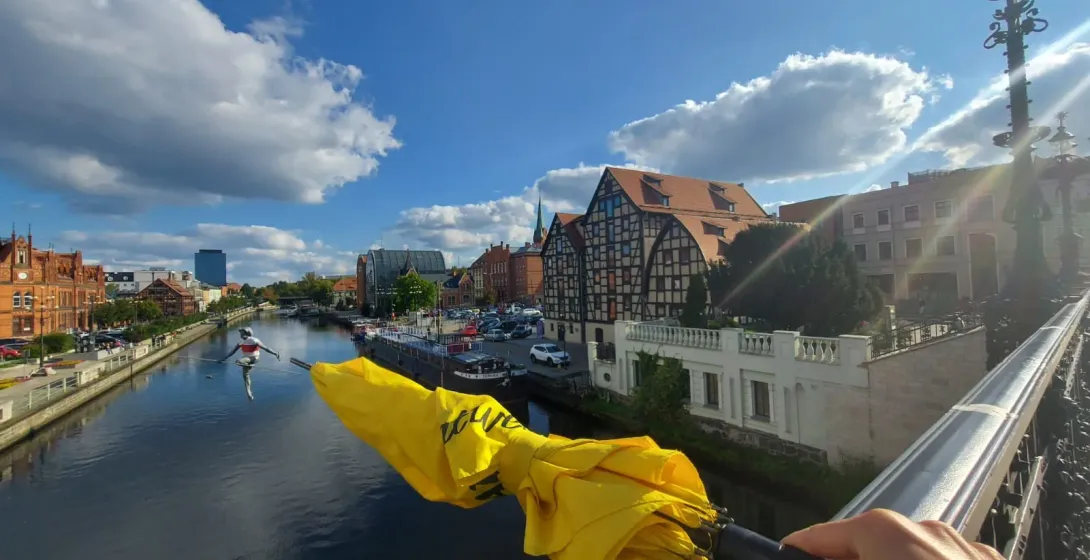 Żółty parasol trzymany na moście z widokiem na rzekę z zabytkowymi budynkami i statuetką chodźca po linie.