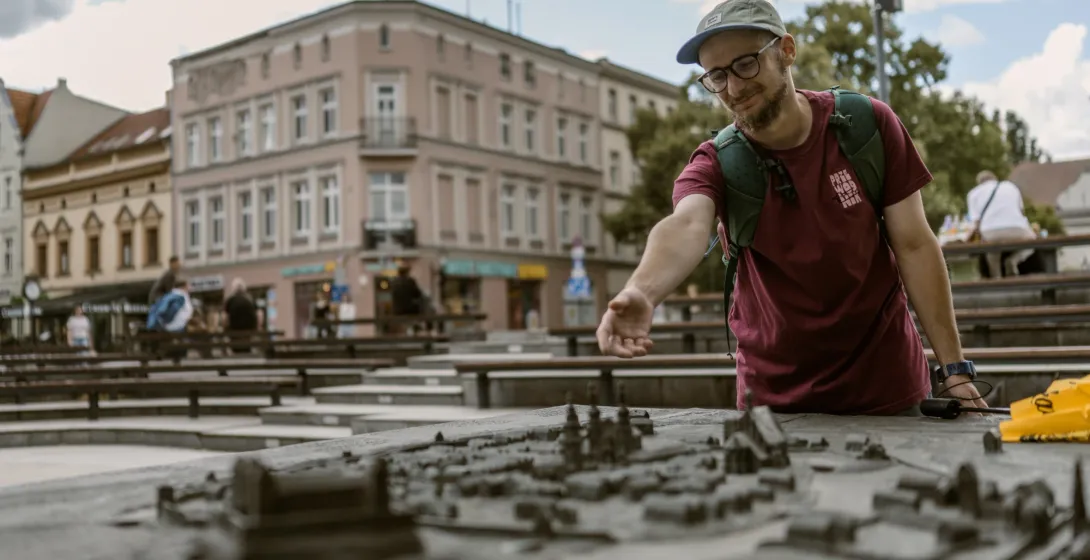 Mężczyzna w czapce i okularach ogląda brązową makietę Starego Miasta, w tle widać kamienice.