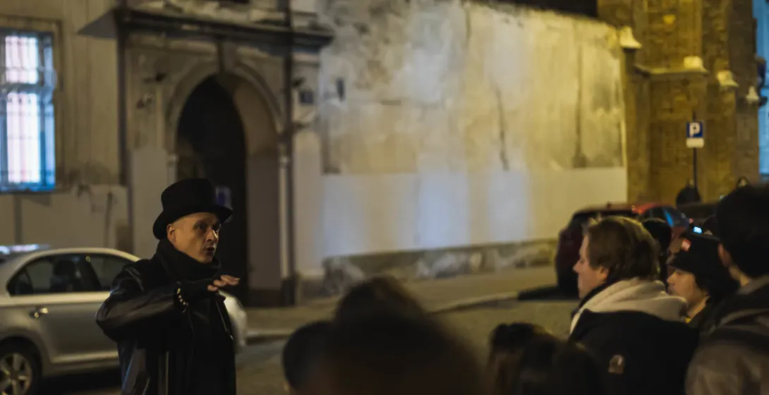 Tour guide in top hat and long coat speaking to a group at night on a cobblestone street in Krakow. Historic buildings are...
