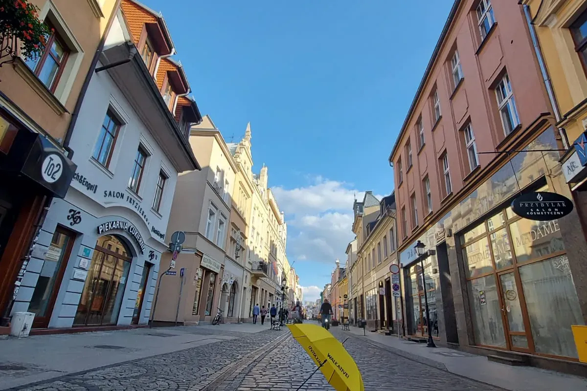 Widok z niskiej perspektywy na brukowaną ulicę w Bydgoszczy z zabytkowymi kamienicami i żółtym parasolem Walkative na pier...