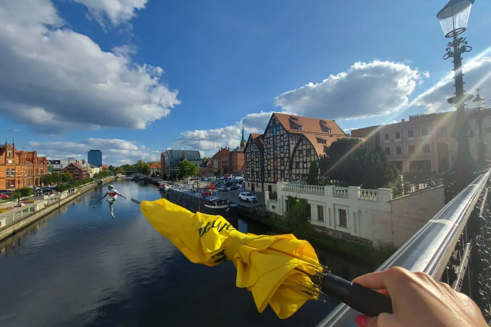 Dłoń trzymająca żółty parasol nad rzeką Brdą, rzeźba linoskoczka, zabytkowe budynki i niebieskie niebo z chmurami.