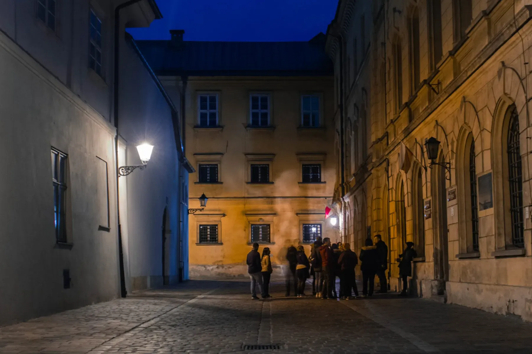 A tour group stands on a narrow, lamplit cobblestone street in Krakow at night, surrounded by historic buildings.