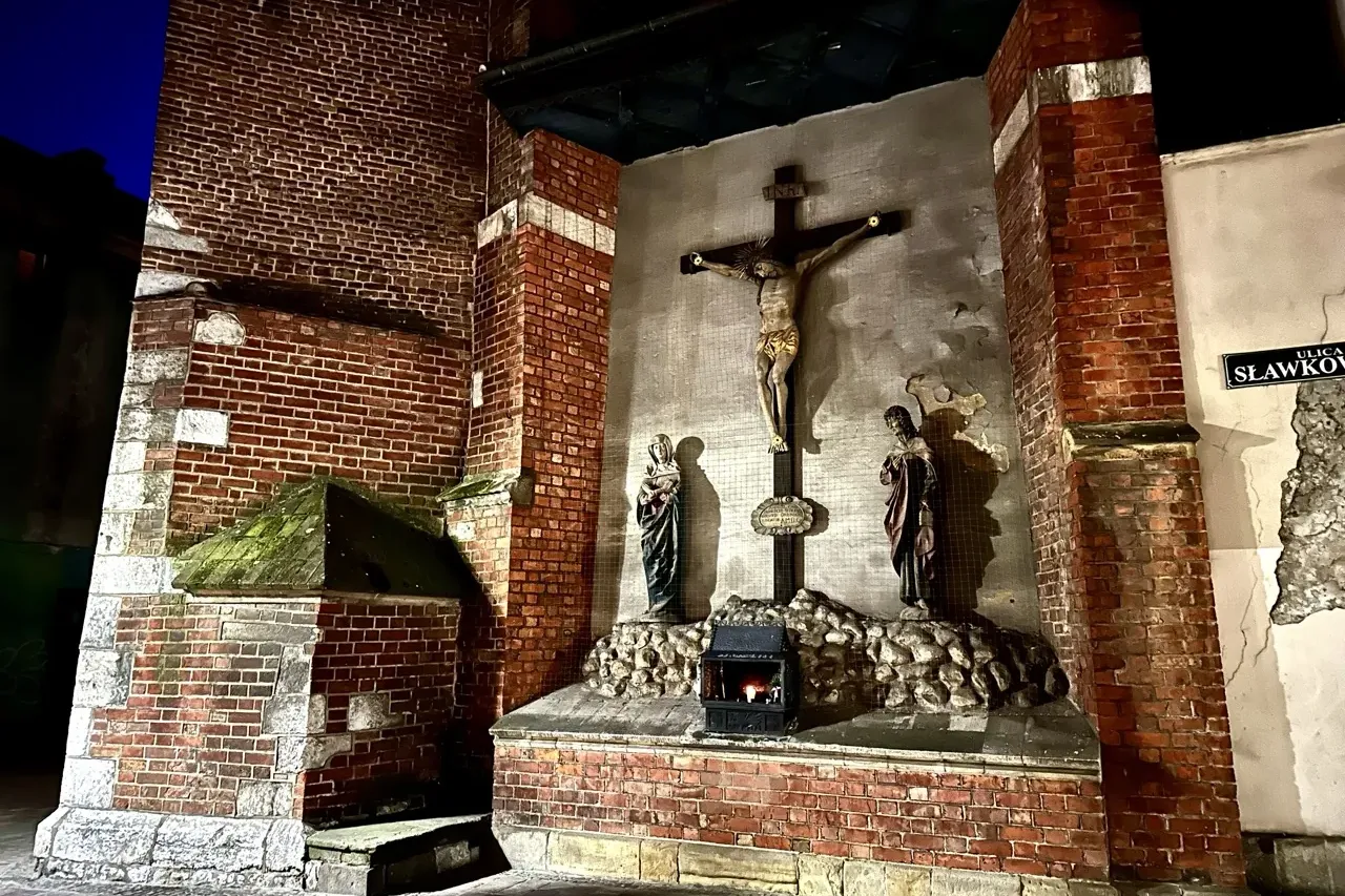 A streetside shrine with a crucifix set into a brick wall on Sławkowska Street in Krakow at night.
