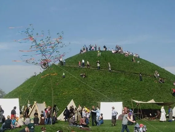 Large green Krakus Mound with many people on its slopes and around tents at the base, under a clear blue sky. A tall pole wit