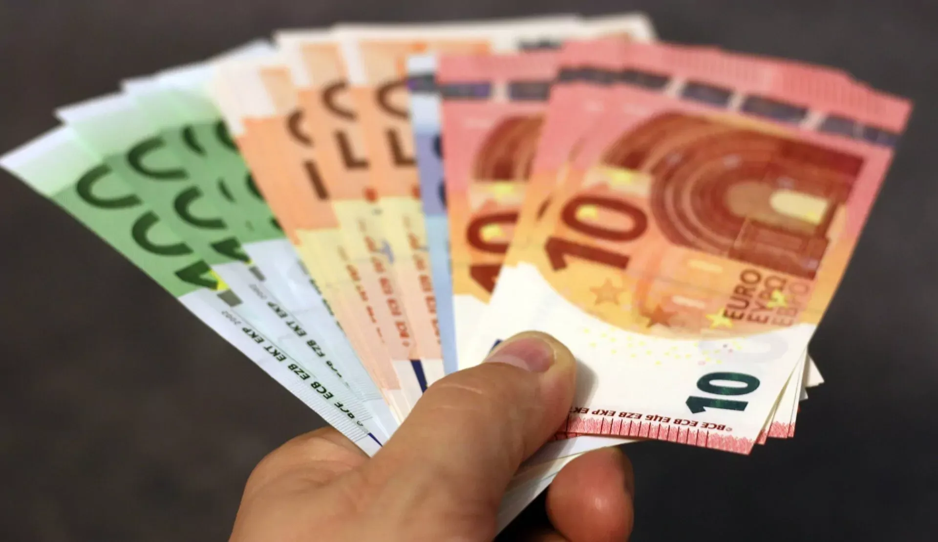 Ręka trzyma wachlarz różnych banknotów euro, w tym nominały 10, 20, 50 i 100, na ciemnym tle.