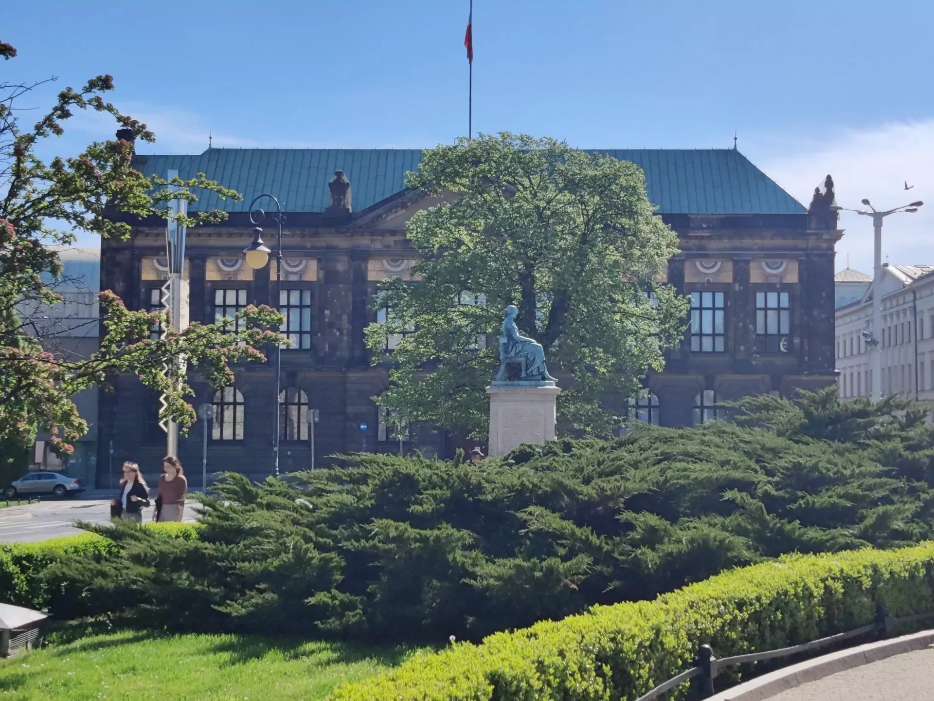 View of the National Museum in Poznań with a seated statue in the foreground, surrounded by lush green bushes and trees.