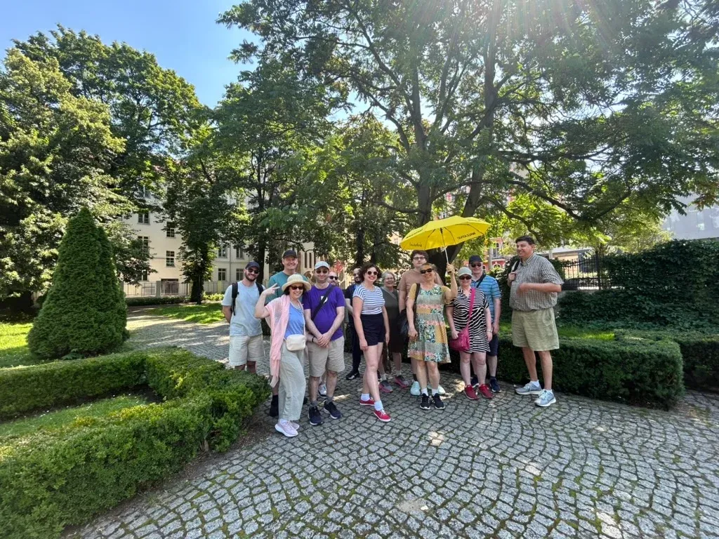 A group of people listening to a male guide with a yellow umbrella and microphone on a cobblestone path in a green park.