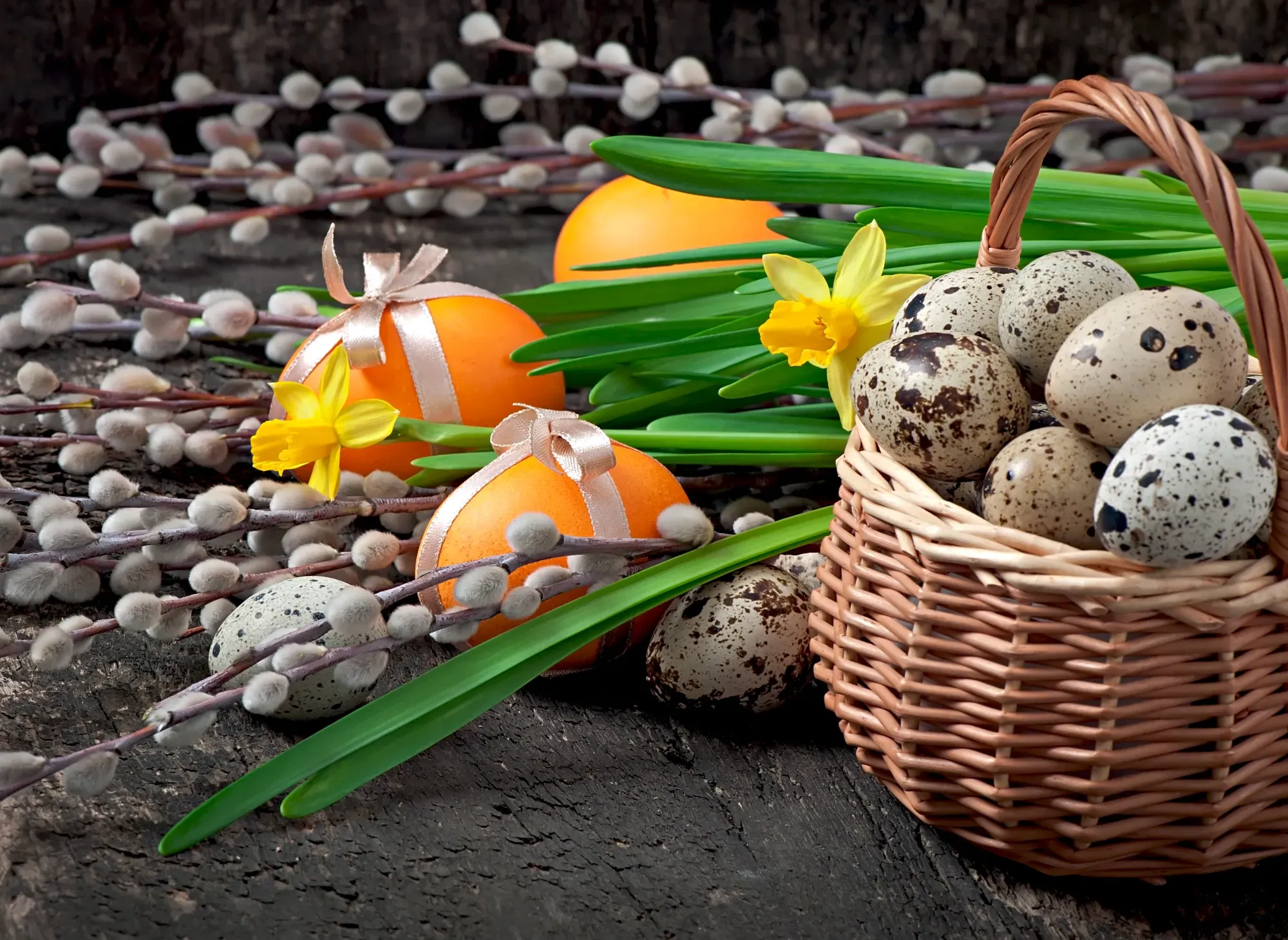 Pussy willow branches, orange eggs with ribbons, yellow daffodils, and a woven basket of speckled eggs on dark wood.