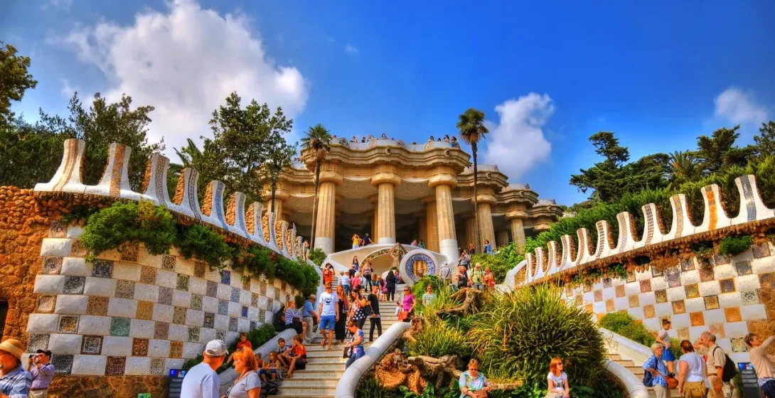 Mosaic-tiled walls and steps lead up to the Hypostyle Room in Park Güell, Barcelona, with numerous tourists on a sunny day.