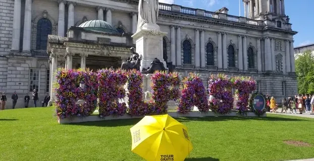 Floral BELFAST sign in front of City Hall; yellow umbrella suggests a free walking tour.