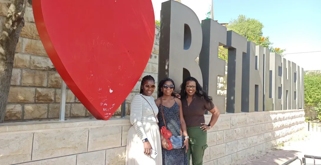 Three women pose happily in Bethlehem, Palestine, in front of a large red heart and the city's name.