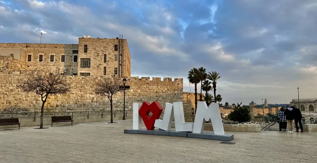 Tourists visit the iconic "I ♥ JLM" sign in Jerusalem, with the Old City walls in the background.