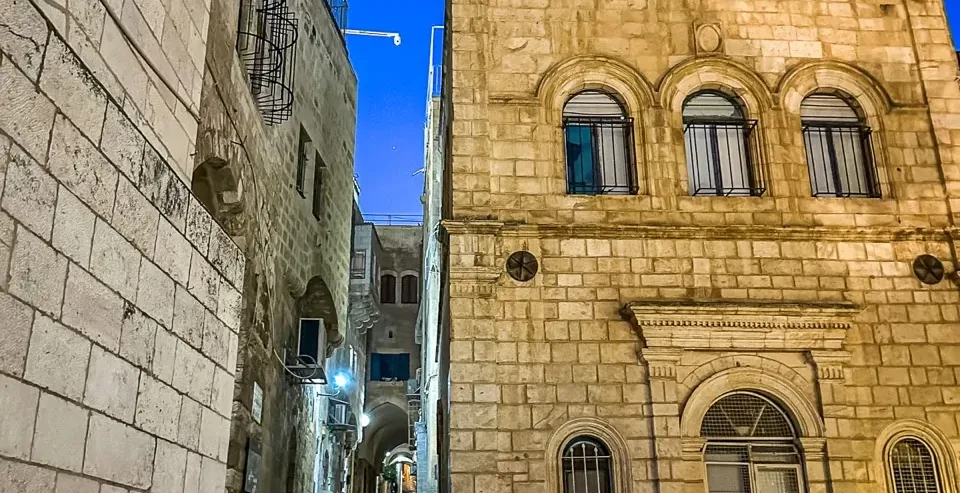 A person walks down a stone staircase in the illuminated Old City of Jerusalem at night.