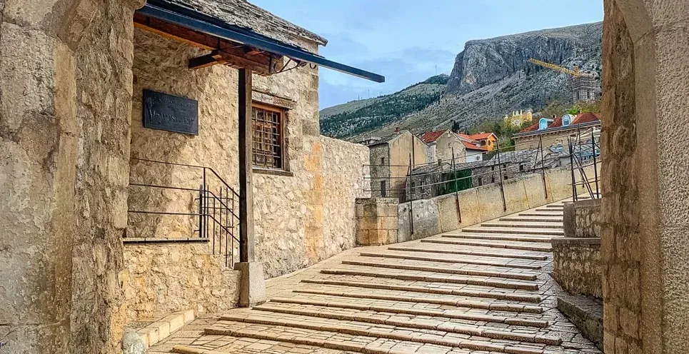 Stone archway framing a staircase leading to a historic building with a mountain backdrop in Mostar.