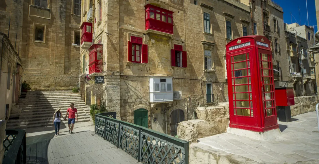 Couple walking across a bridge in Valletta, Malta, with historic buildings and a red telephone booth.