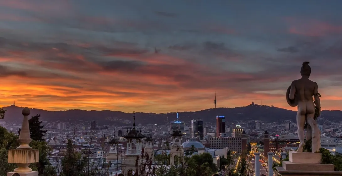 View of Barcelona city skyline at sunset with a statue in the foreground and Tibidabo mountain in the background.