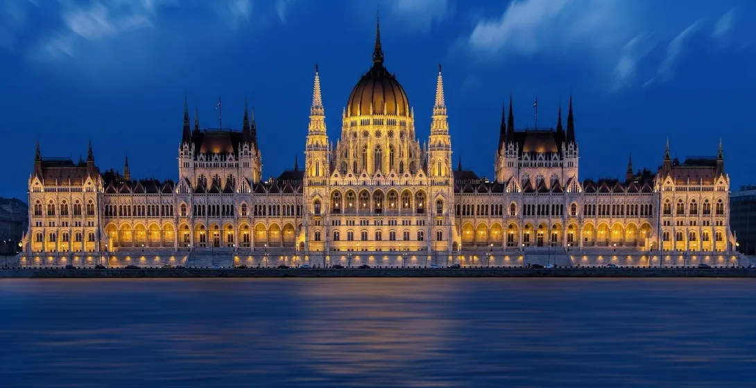 The illuminated Hungarian Parliament Building at night, viewed across the dark blue Danube River under a cloudy sky.