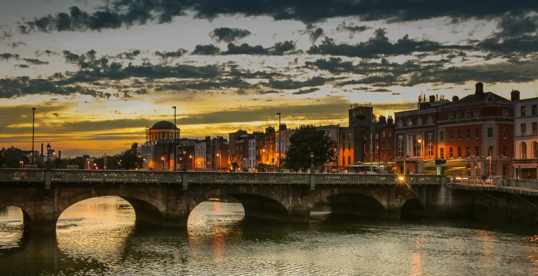 Stone arch bridge over a river in Dublin at sunset, with city buildings lit up on the far bank and a cloudy sky.