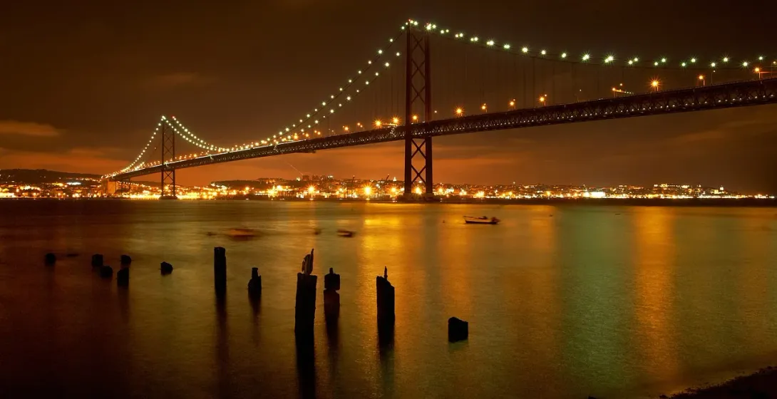 25 de Abril Bridge spanning a river at night, illuminated with city lights reflecting on the water. Wooden posts in foregroun