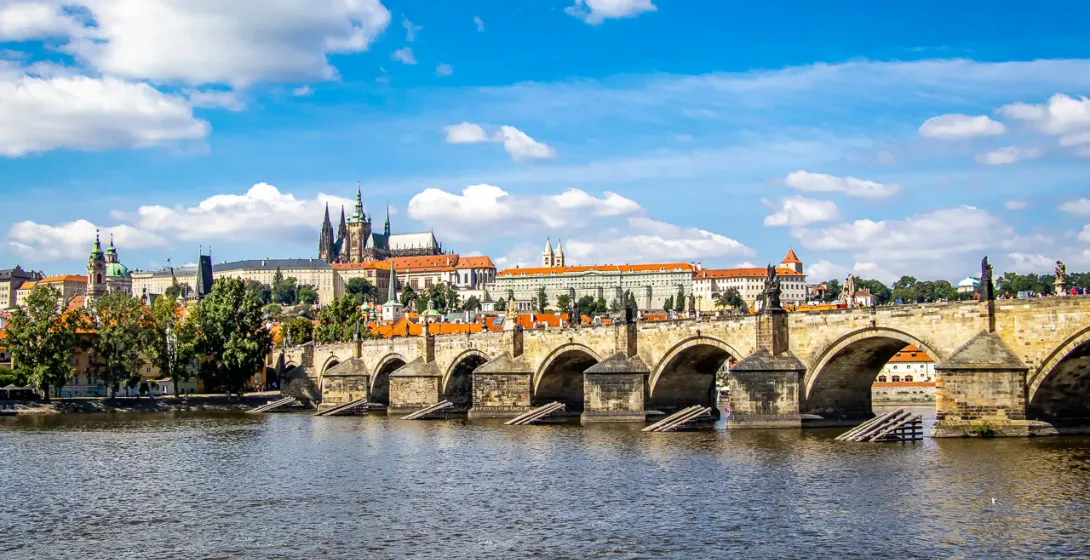 View of Charles Bridge with statues and arches spanning a river, Prague Castle on the distant left bank under a blue sky with