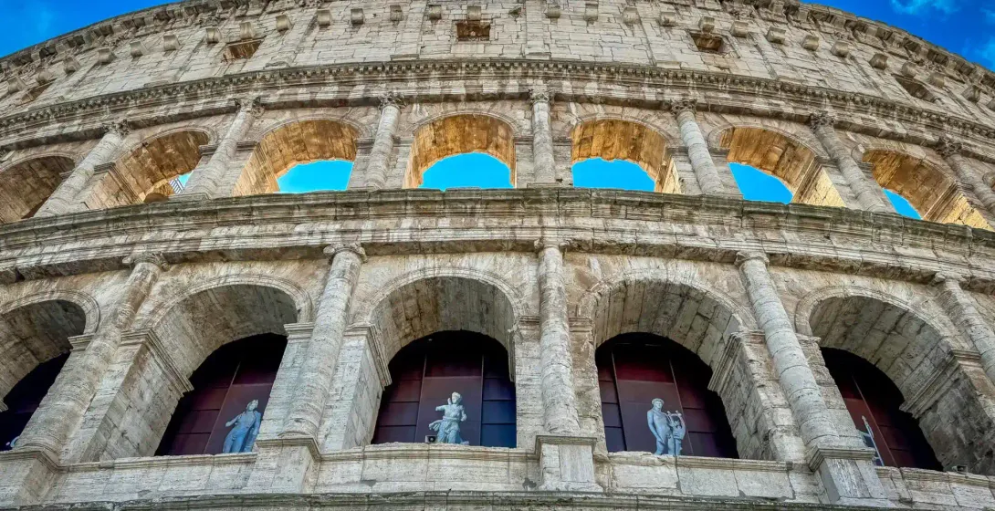 Exterior view of the Colosseum in Rome, showing multiple tiers of stone arches, columns, and statues in lower arches, against