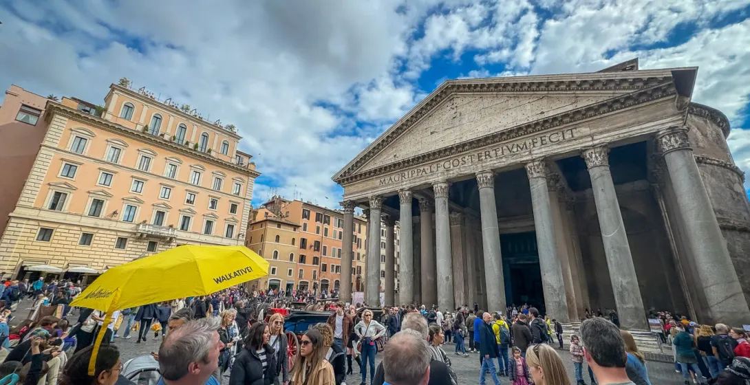 Crowded square in front of the Pantheon in Rome, showing its large portico with columns and an ancient inscription. A yellow
