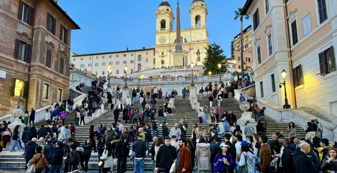 Crowds of people gather on the Spanish Steps in Rome at dusk, leading up to the Church of Trinità dei Monti with an obelisk.