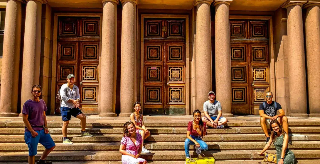 Nine travelers sitting and standing on stone steps in front of a grand building with tall columns and three ornate wooden doo