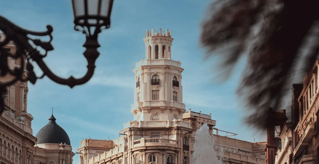 Historic buildings in Valencia, including a prominent tower, seen from a street. A large fountain sprays water, with ornate s