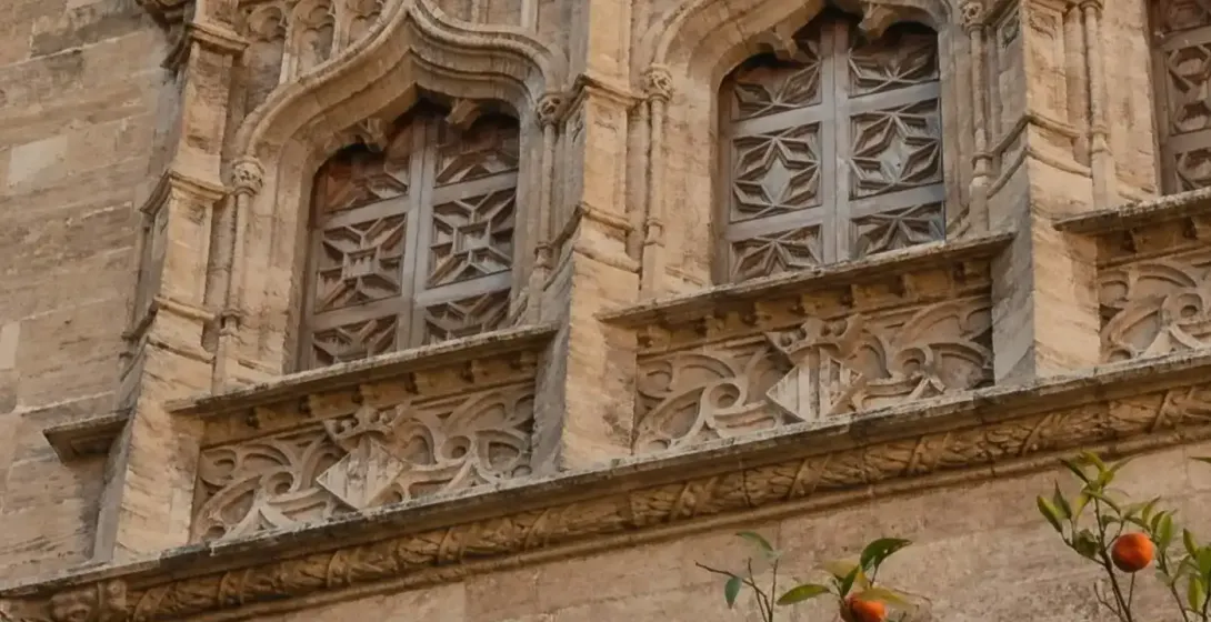 Ornate stone building with gothic windows, carved details, and a flag, partially covered by a tree full of ripe oranges.