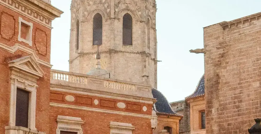 Historic brick buildings and a bell tower in a sunny European square with a statue and orange trees.