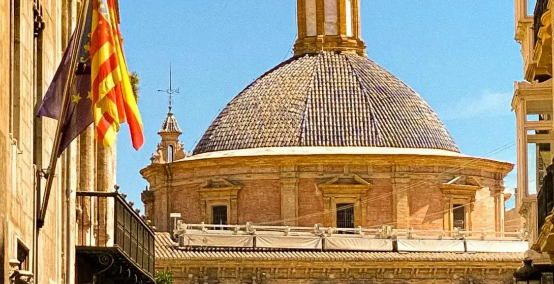 Narrow pedestrian street in Valencia with people walking, leading towards a prominent church building with a tiled dome and l