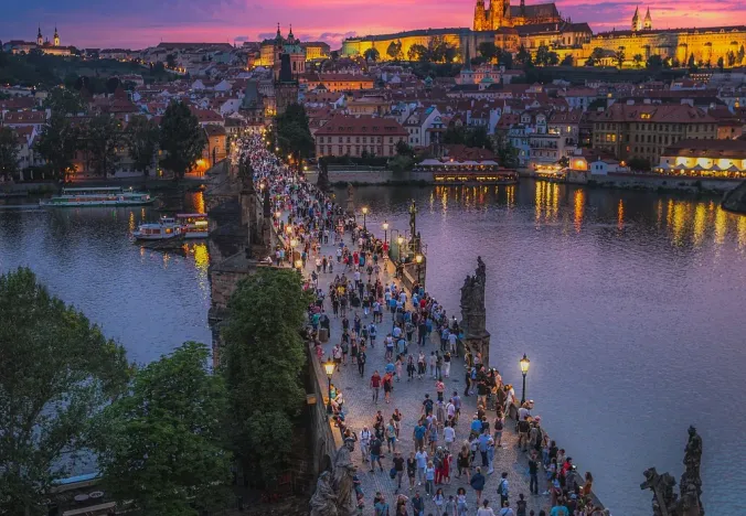 Tourists stroll across Charles Bridge in Prague at sunset, with Prague Castle majestically lit in the background.