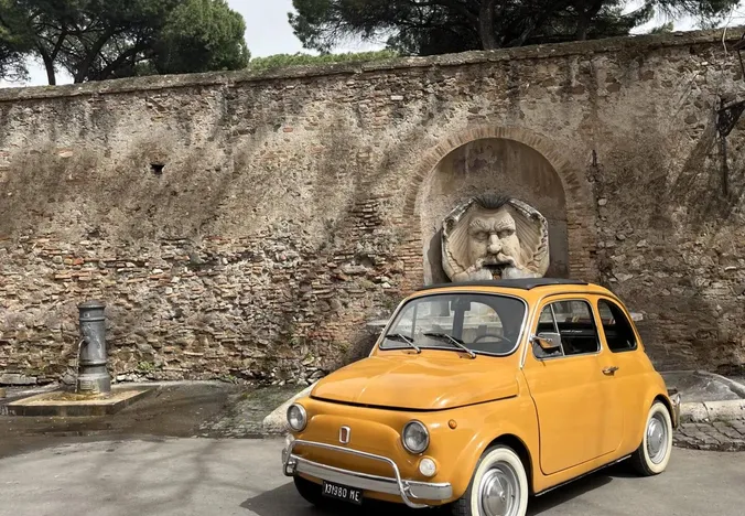 A yellow Fiat 500 parked near a Roman wall with a unique fountain.