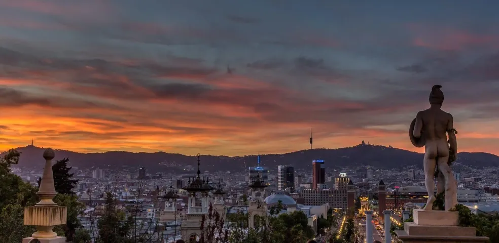 Panoramic view of Barcelona at sunset, with a statue in the foreground and Tibidabo Mountain in the distance.