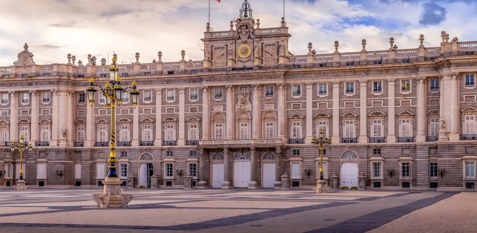 Grand Royal Palace of Madrid with a Spanish flag on top, two ornate street lamps, and a wide empty plaza under a dramatic clo