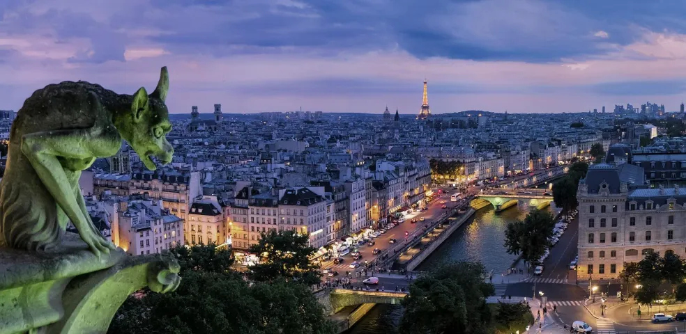 Stone gargoyle on a building overlooking the Paris cityscape at dusk, with the illuminated Eiffel Tower and city lights.