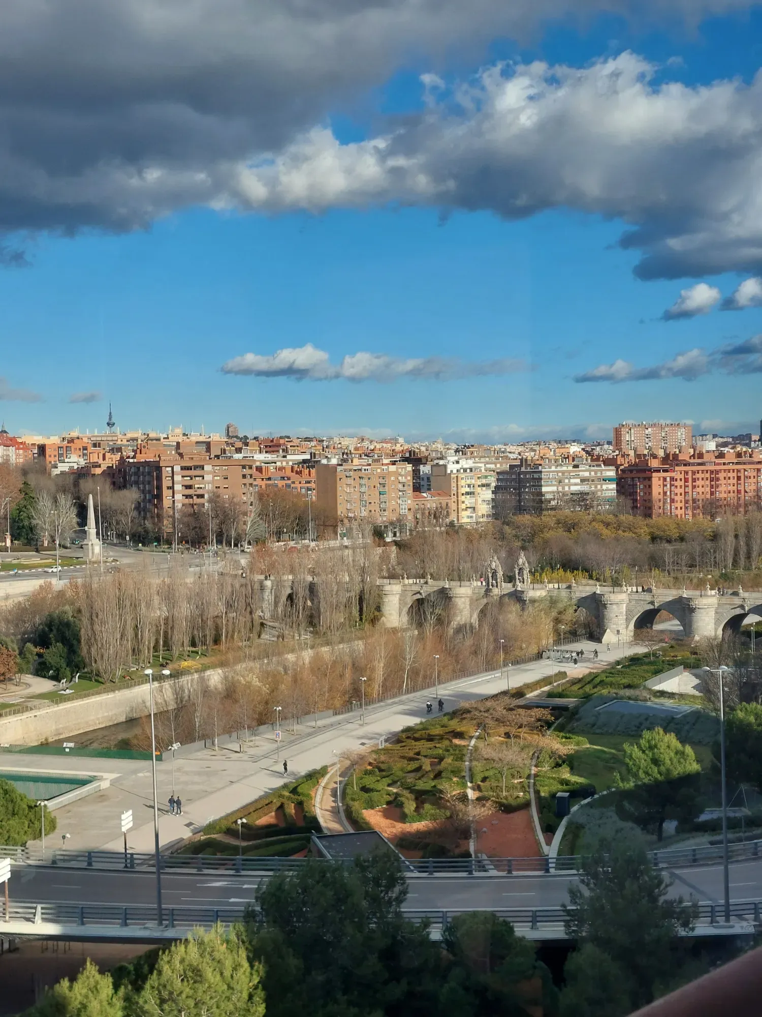 View over the Manzanares River in Madrid, with the historic Puente de Toledo bridge and city buildings under a partly clou...