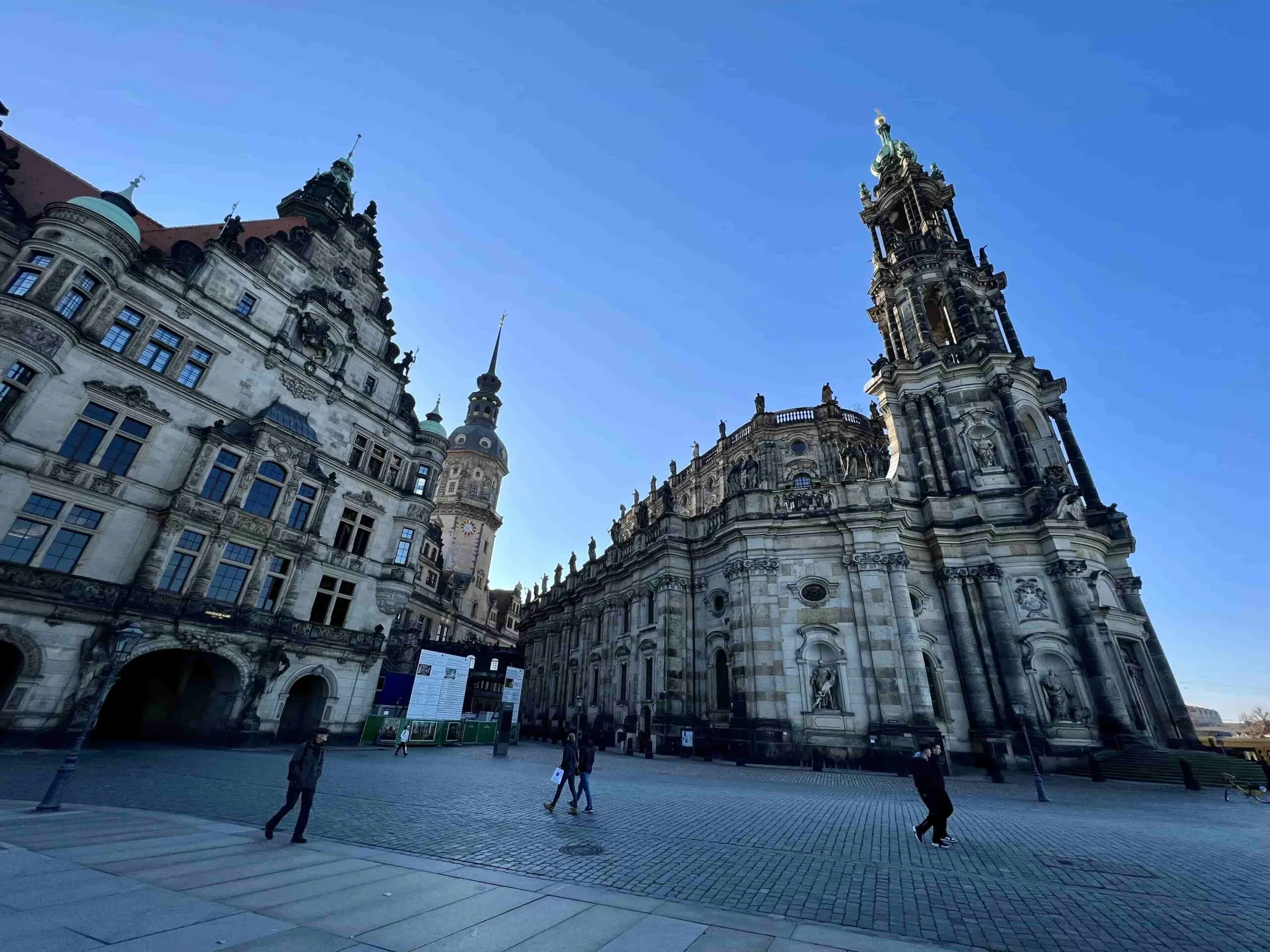 Historic Dresden Royal Palace and Hofkirche under a clear blue sky, with people walking on the cobblestone square.