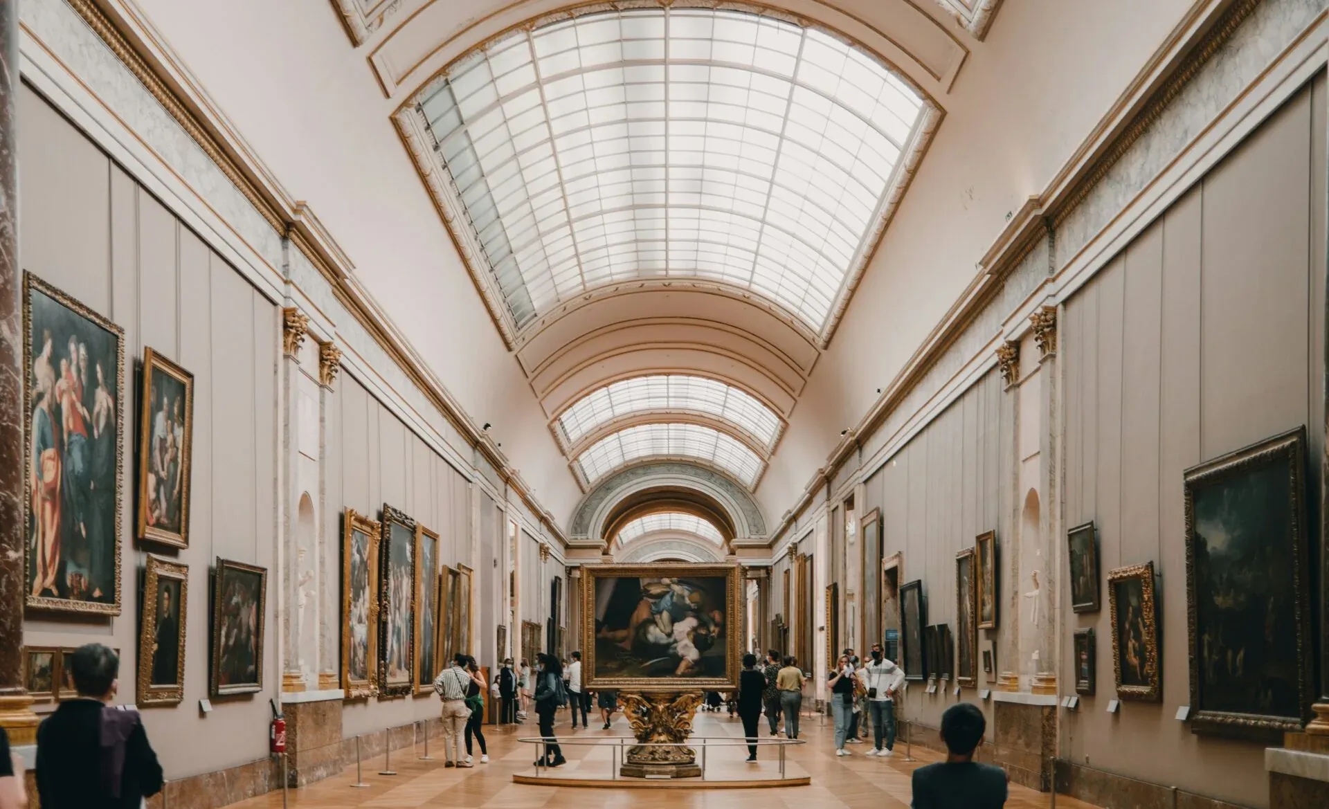 Grand art gallery in Paris with numerous classical paintings lining the walls and a large vaulted skylight ceiling. Visitors