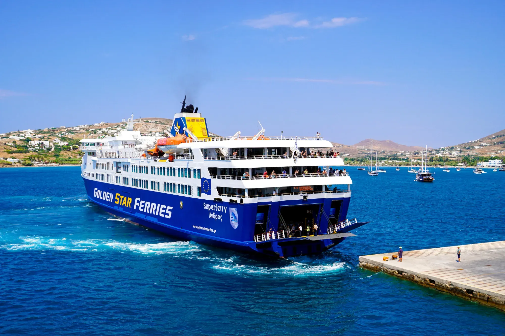 Blue and white Golden Star Ferries passenger ship approaching a concrete pier on a Greek island.