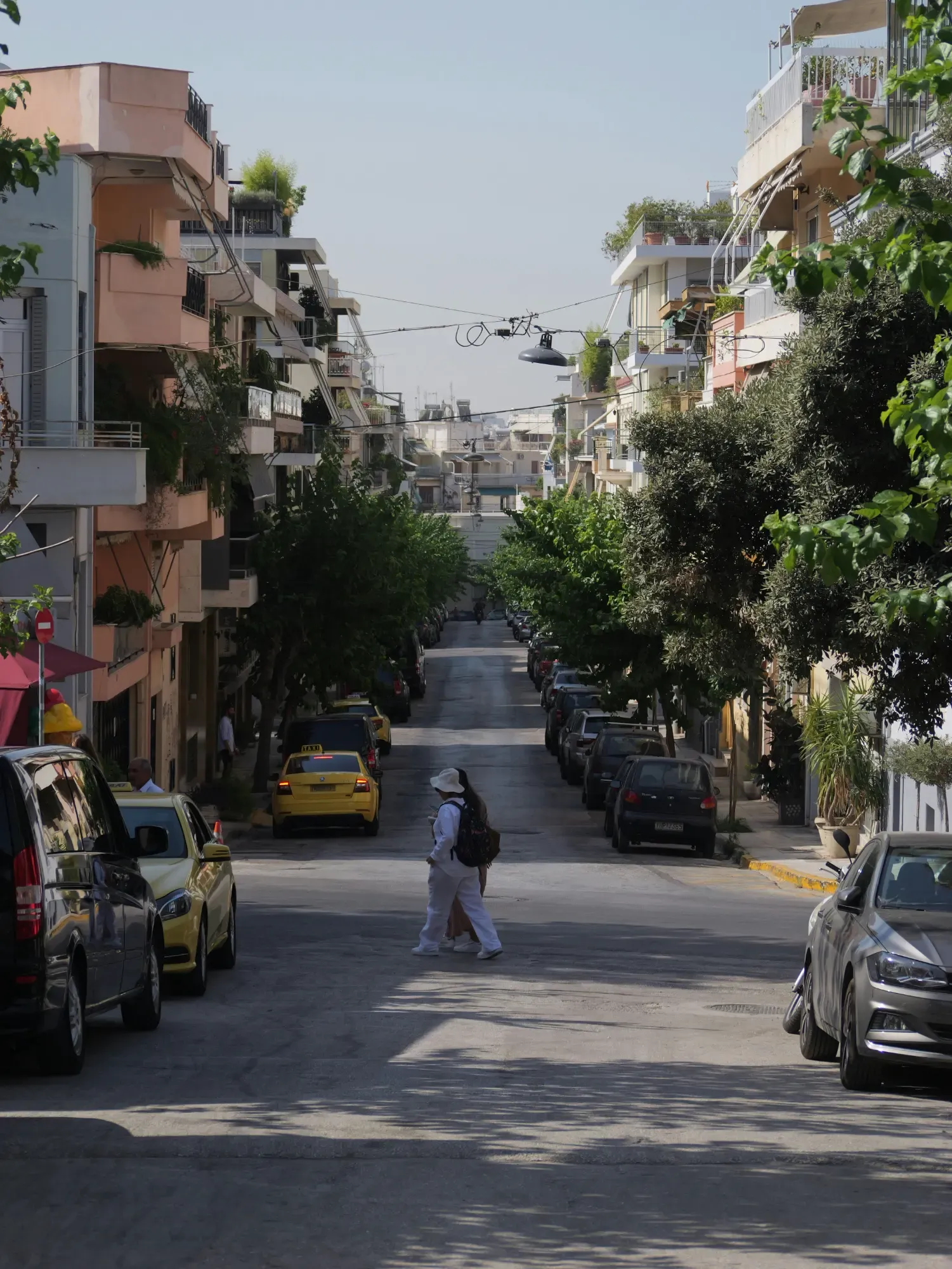 View down a residential street in Koukaki, Athens, lined with apartment buildings, parked cars, and trees.