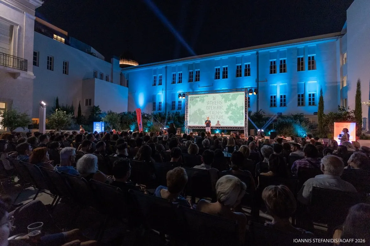 A crowd watches a speaker on stage at the Athens Open Air Film Festival at night, in a courtyard with blue lighting.