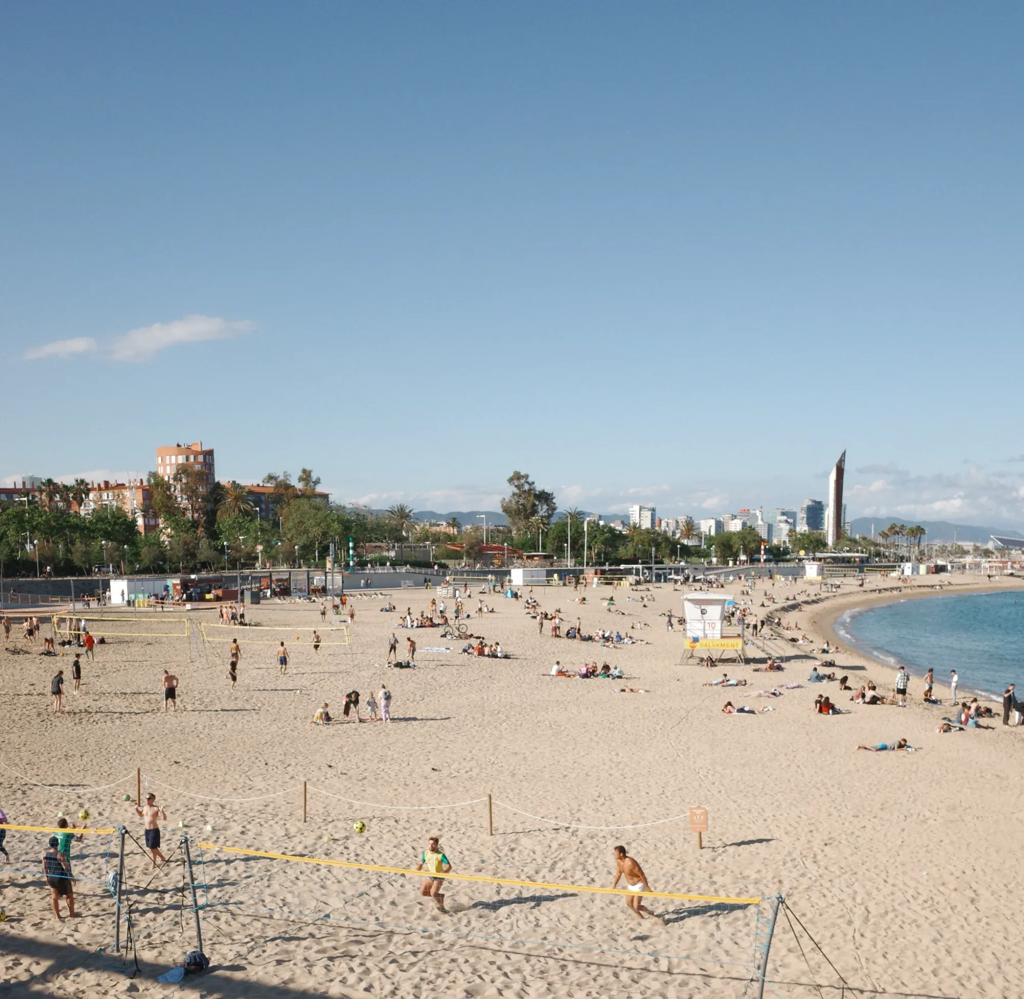 A sunny day at a crowded Barcelona beach, with people playing volleyball and relaxing on the sand.