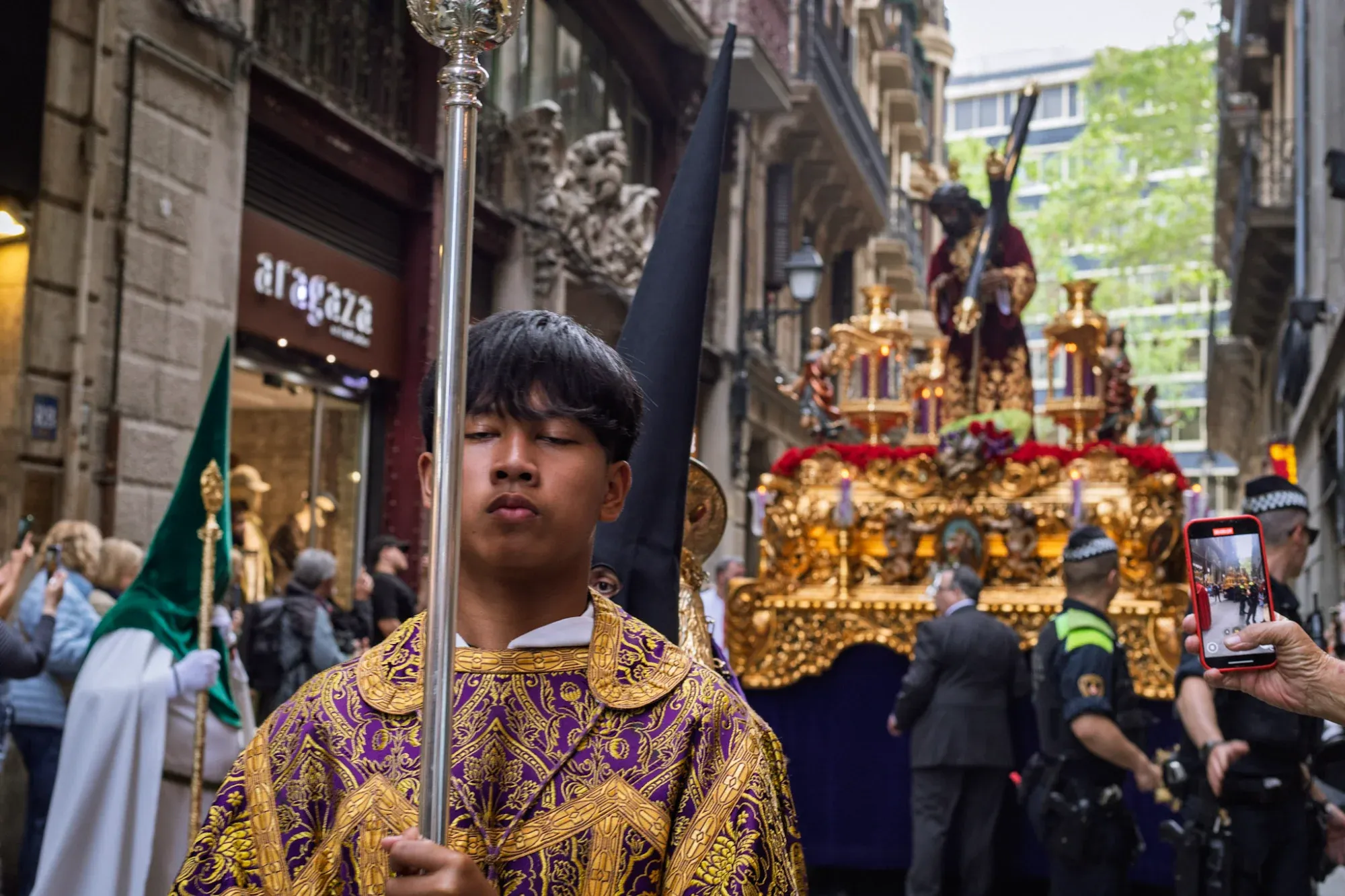 Young person in purple vestments leading a Holy Week procession in Barcelona, with a golden float in the background.