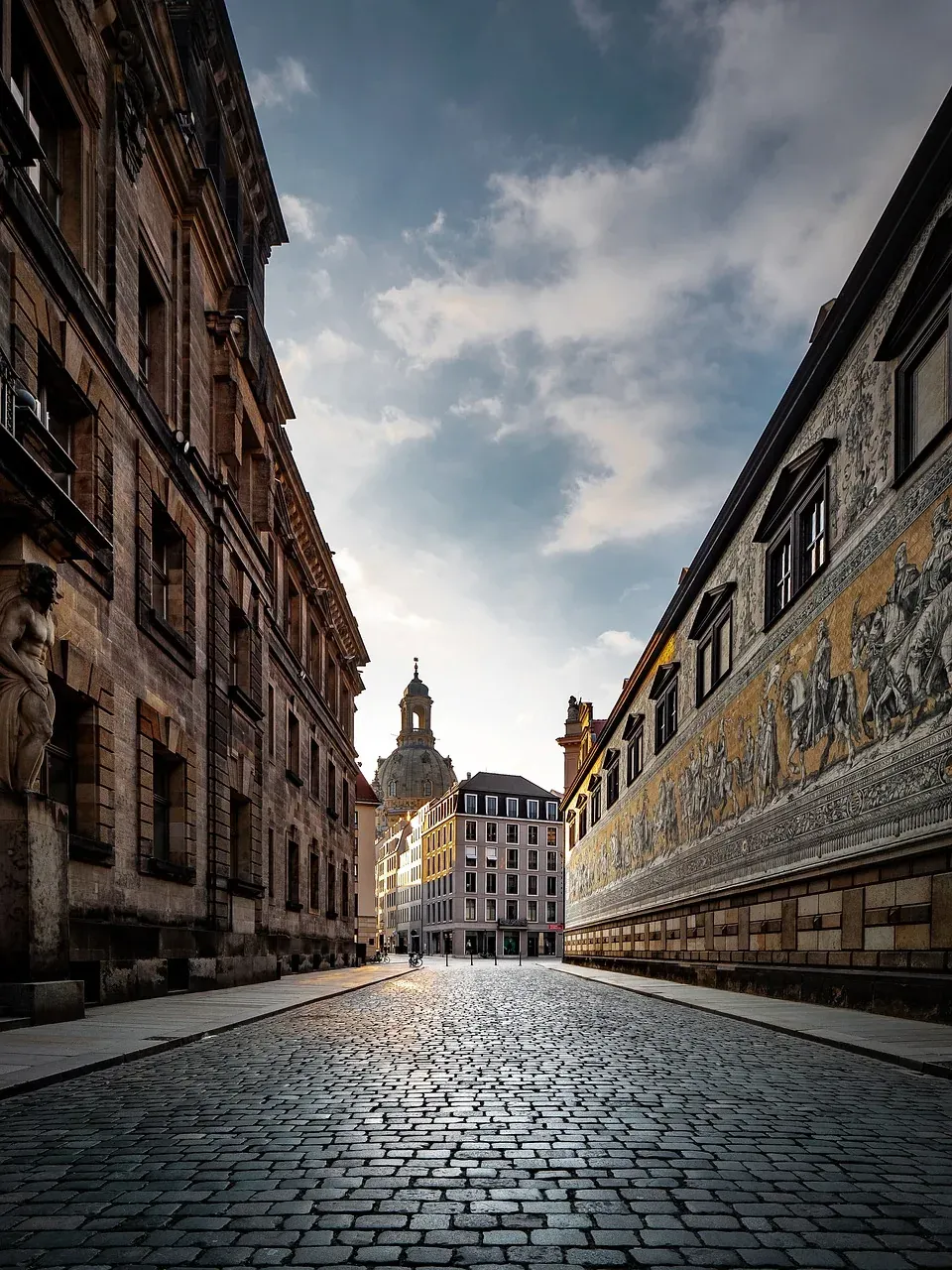 Cobblestone street in Dresden leading to Frauenkirche dome, flanked by old buildings and the Fürstenzug mural on the right.