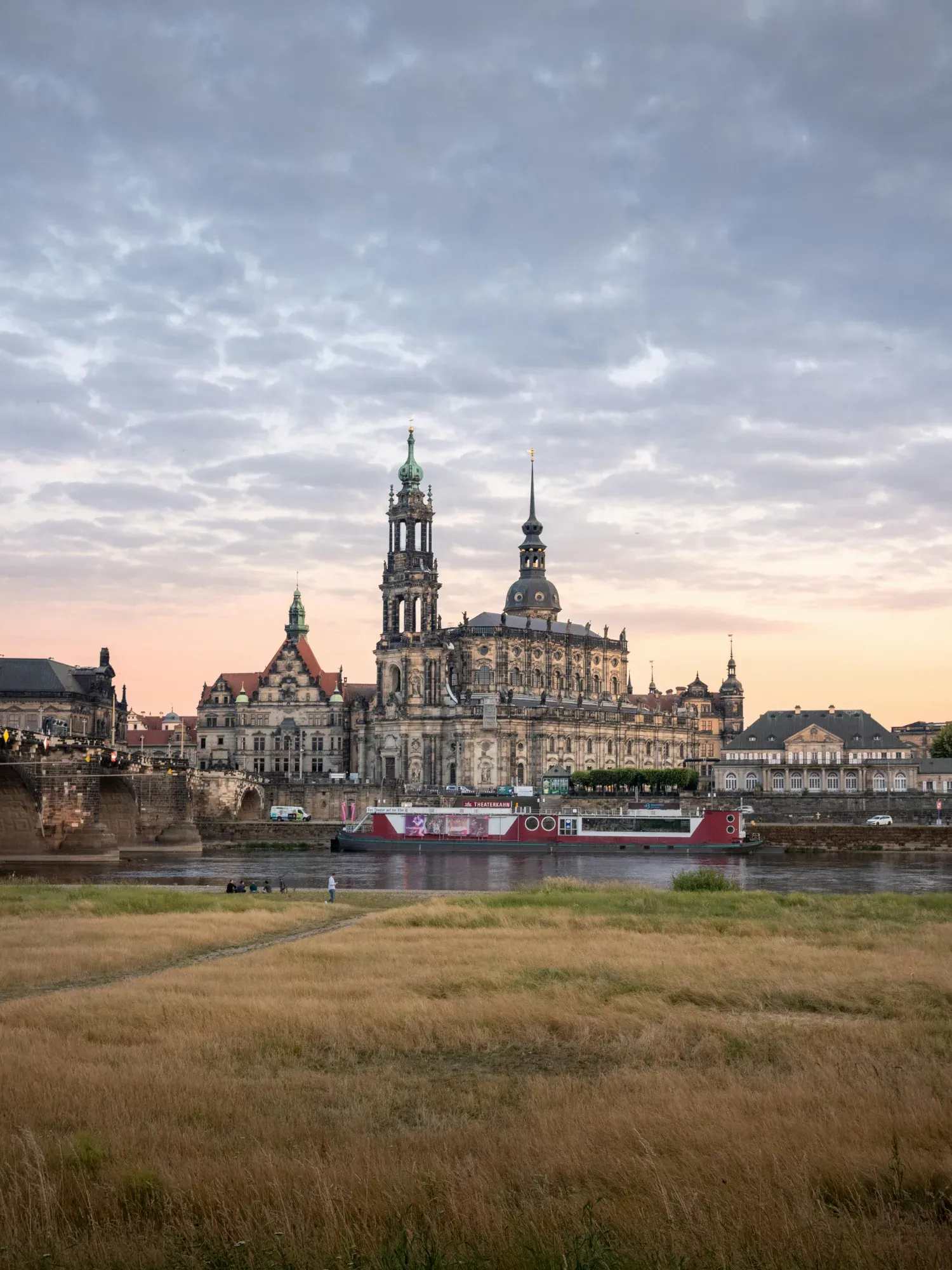 Dresden skyline with the Cathedral and Elbe river viewed from a grassy bank at dusk.