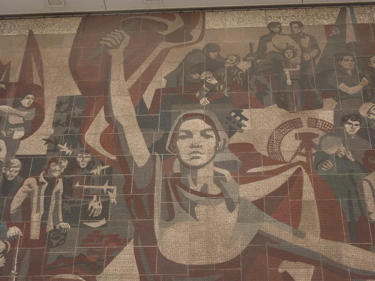 A large socialist-realist mosaic mural in shades of red, grey, and beige, depicting workers, flags, and a central figure w...